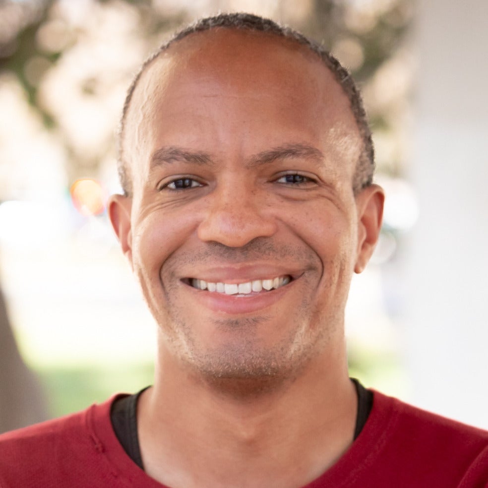 Andrew Lewis's headshot. A person with a buzzed haircut wearing a long-sleeved shirt is outside with their arms crossed, smiling at the camera.