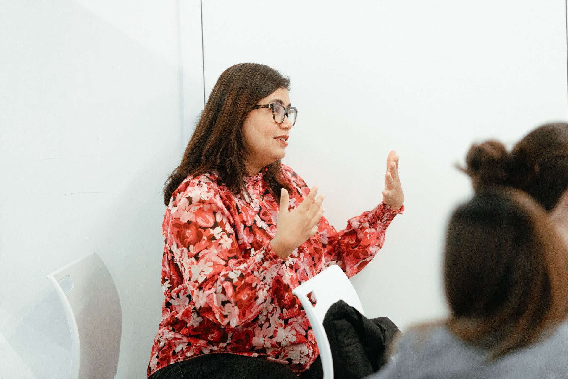 A woman with shoulder-length straight brown hair, glasses, and a pink floral long sleeve shirt sits against a white backdrop. She is speaking and gesturing with her hands.